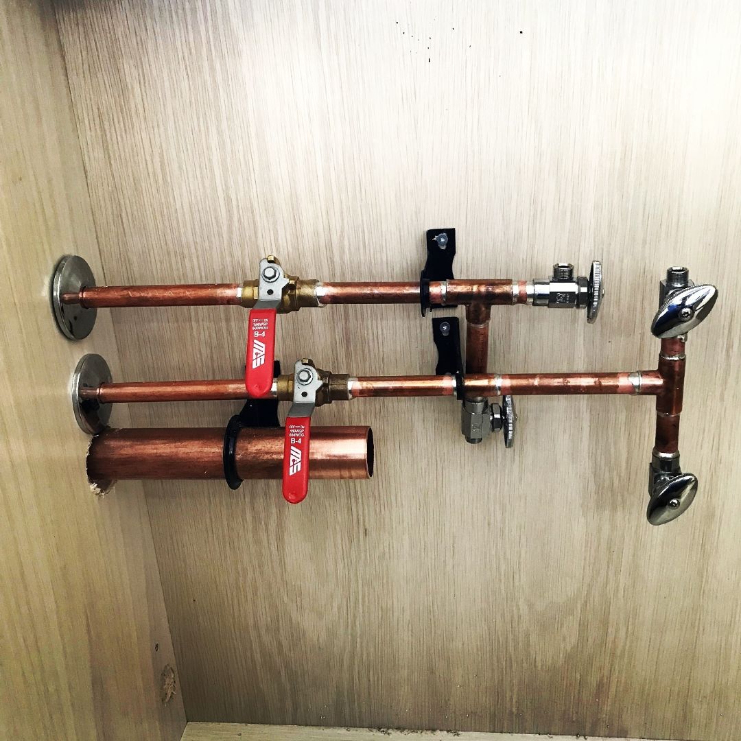 Two parallel copper water supply pipes mounted on a light wood wall inside a cabinet. Each pipe has a red-handled ball valve, and they are secured with black pipe straps. The pipes connect to chrome shut-off valves at the ends, likely for a sink or vanity installation.