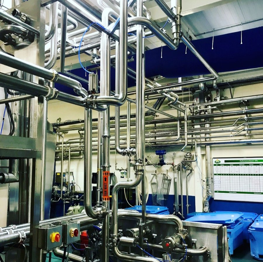 Complex stainless steel industrial piping system inside a manufacturing or processing facility, featuring valves, control panels, and overhead pipe routing.