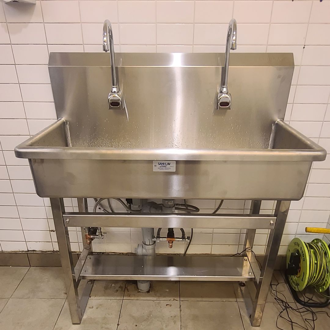 A commercial stainless steel double faucet hand wash station with a deep basin and backsplash, mounted against a tiled wall. Plumbing and water supply lines are visible beneath the sink, along with a floor-mounted hose reel on the right side.