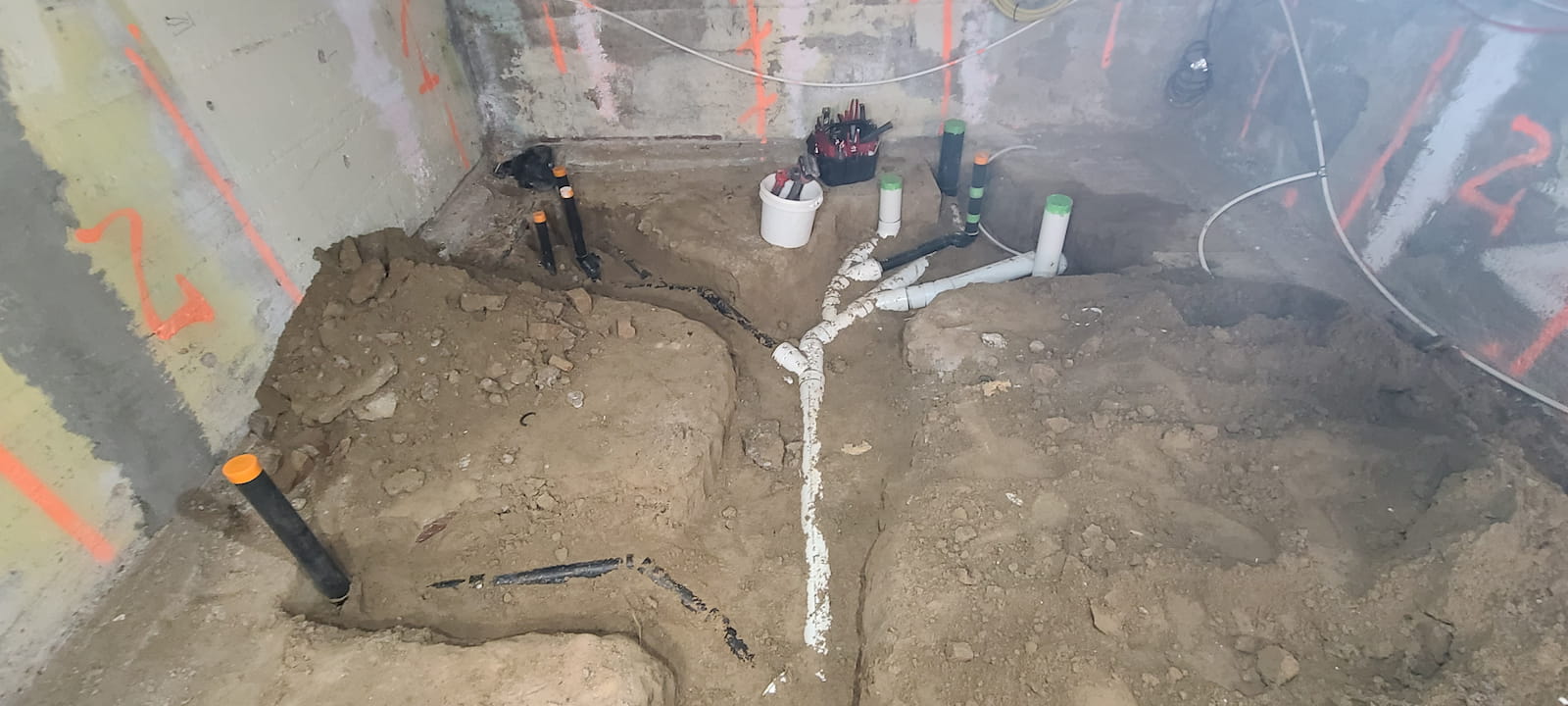 Basement construction site with exposed rough-in plumbing for drains and vent pipes, surrounded by dirt trenches and utility markings on concrete walls.