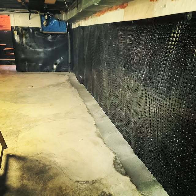 Basement wall waterproofing installation with black dimpled drainage membrane applied along the perimeter, over a concrete foundation, with a clean concrete floor in the foreground.