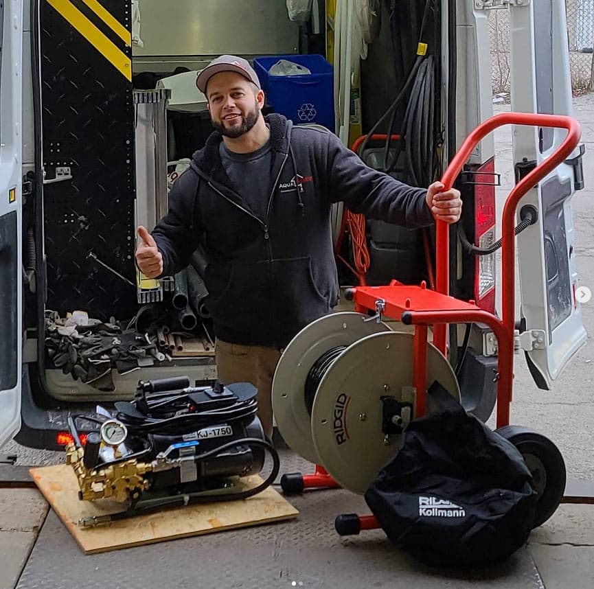 A person in work attire stands beside specialized plumbing equipment, including a cable reel and jetter, in front of an open service van loaded with tools and supplies.