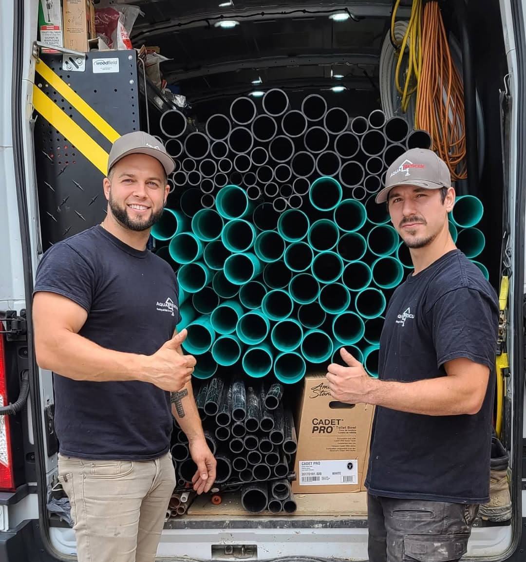 Two men wearing gray caps and black Aqua Rescue t-shirts stand in front of the open back of a van, giving thumbs up. Behind them, the van is stacked with organized green and black PVC pipes, as well as some boxes and coiled cables. The men are smiling and posing for the photo.