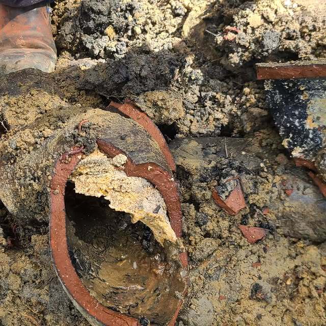 A broken and collapsed terracotta pipe buried in soil, surrounded by dirt, gravel, and small debris.