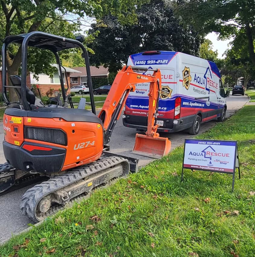 An orange mini excavator is parked on the street beside a branded Aqua Rescue Plumbing and Drain van, with a company yard sign placed on the nearby grass in a residential neighborhood.