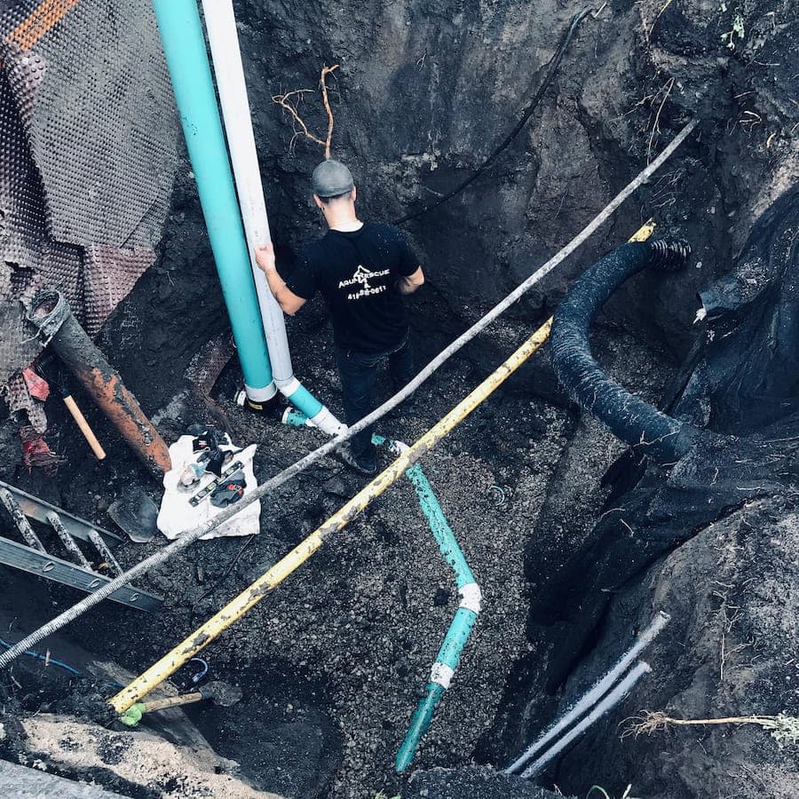 A person stands inside a deep trench working with several exposed green and white pipes, surrounded by dark soil and construction materials.