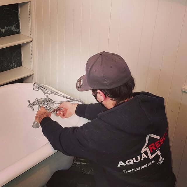 A plumber wearing a dark cap and black "Aqua Rescue Plumbing and Drain" sweatshirt is installing or adjusting a faucet on a white clawfoot bathtub inside a bathroom. The plumber is leaning over the tub, using both hands to work on the fixture.