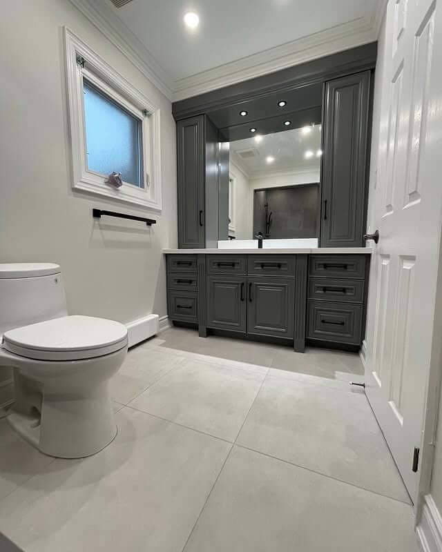 A modern bathroom with a white toilet on the left and a gray vanity with multiple drawers and cabinet doors on the right. Above the vanity is a large mirror with built-in lights, and the floor is covered with large light gray tiles. A frosted window is on the wall above a black towel bar. The overall color scheme is neutral with white walls and gray accents.