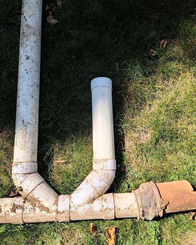 A section of plumbing pipes laid on grass. The setup includes white PVC pipes and an older, discolored clay or terracotta pipe. The pipes form a T-junction, with the PVC pipe extending upward from the joint. Dirt is visible on parts of the pipes.