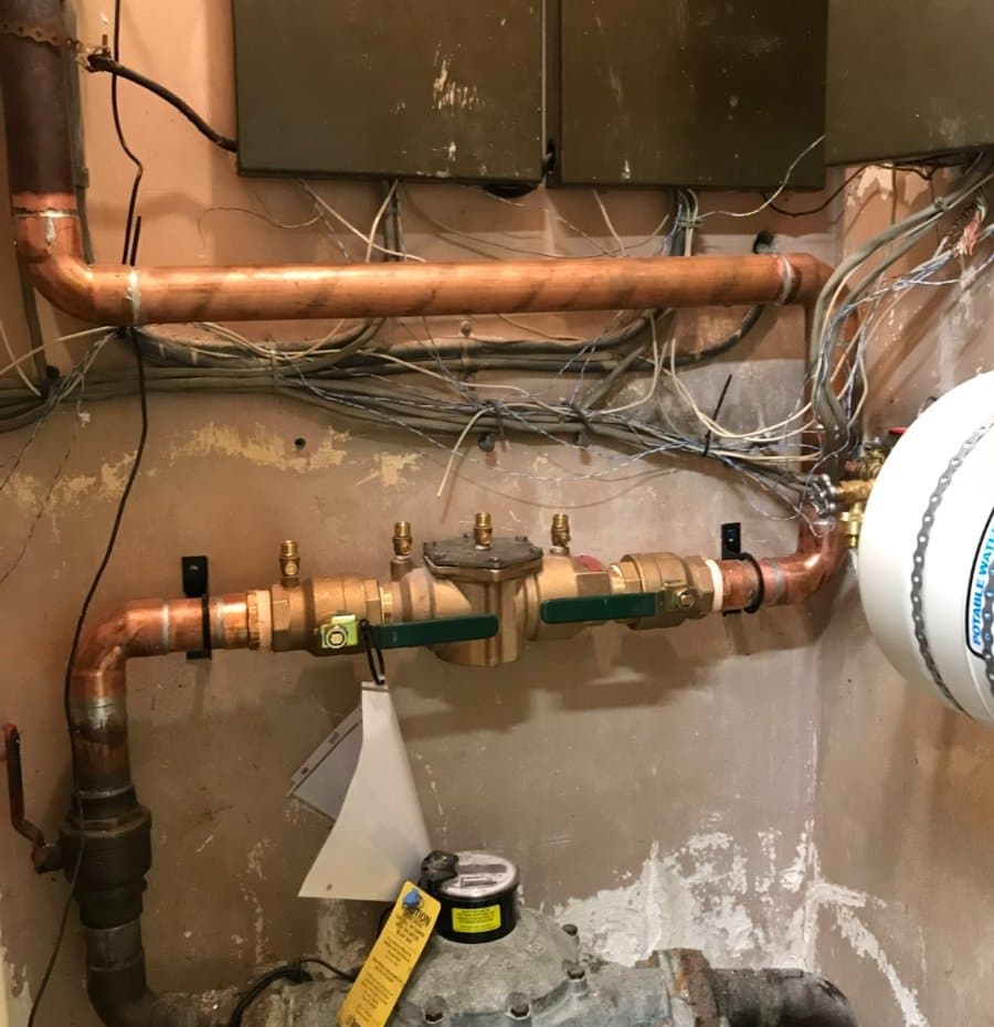 Copper plumbing system with a backflow prevention assembly and shut-off valves installed on an interior utility wall, surrounded by exposed wiring and meters.