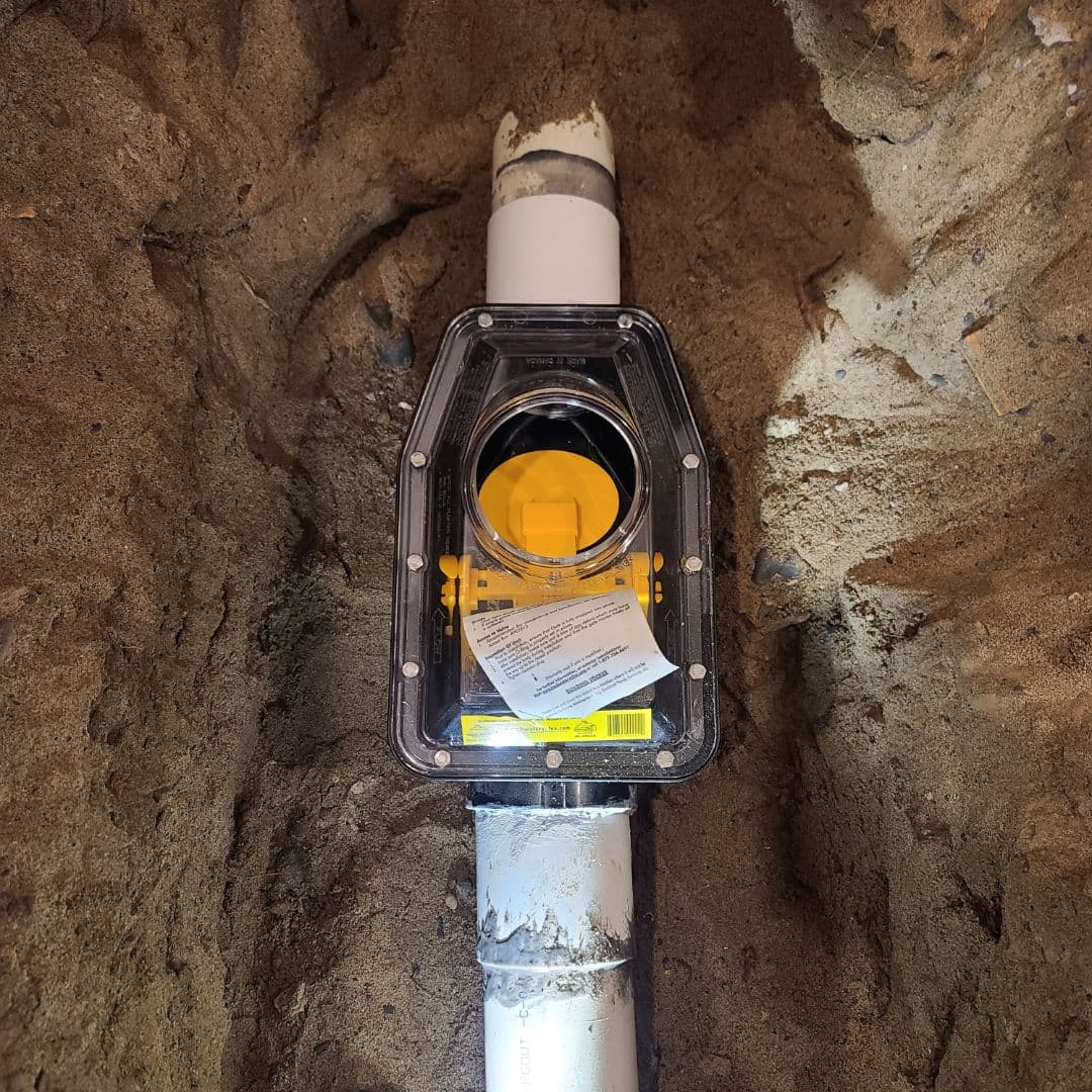 Underground backwater valve installation with clear cover and visible yellow internal flap, connected to white PVC drainage pipe, surrounded by compacted soil in a trench.