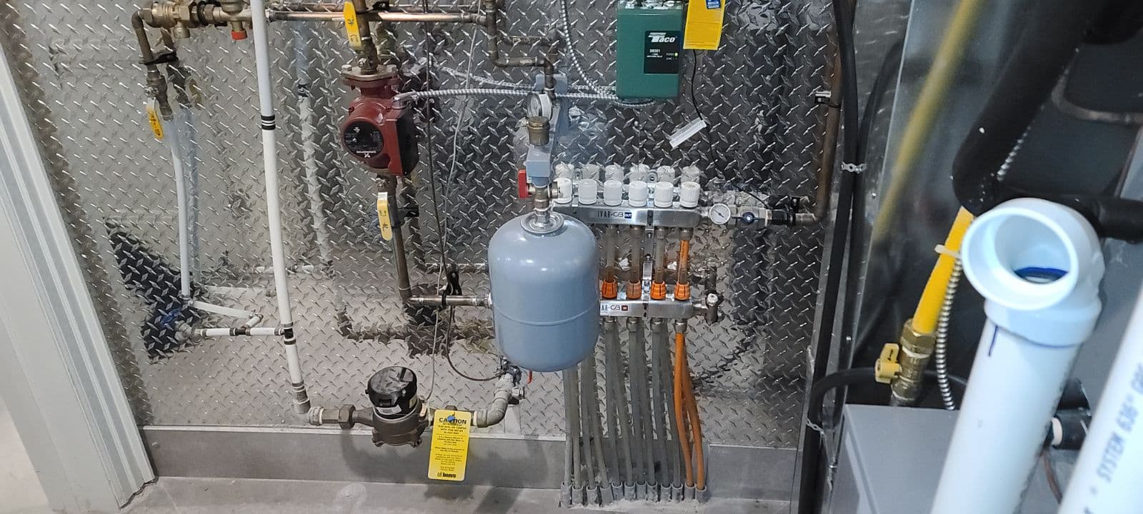 A professionally installed radiant floor heating system setup, featuring a pressurized expansion tank, circulator pump, multiple zone valves, manifold with PEX tubing connections, and control modules mounted on a metallic diamond plate wall. Yellow caution tags are attached to components for safety and maintenance guidance.