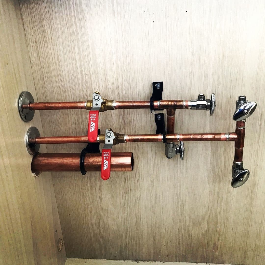 Two parallel copper water supply pipes mounted on a light wood wall inside a cabinet. Each pipe has a red-handled ball valve, and they are secured with black pipe straps. The pipes connect to chrome shut-off valves at the ends, likely for a sink or vanity installation.