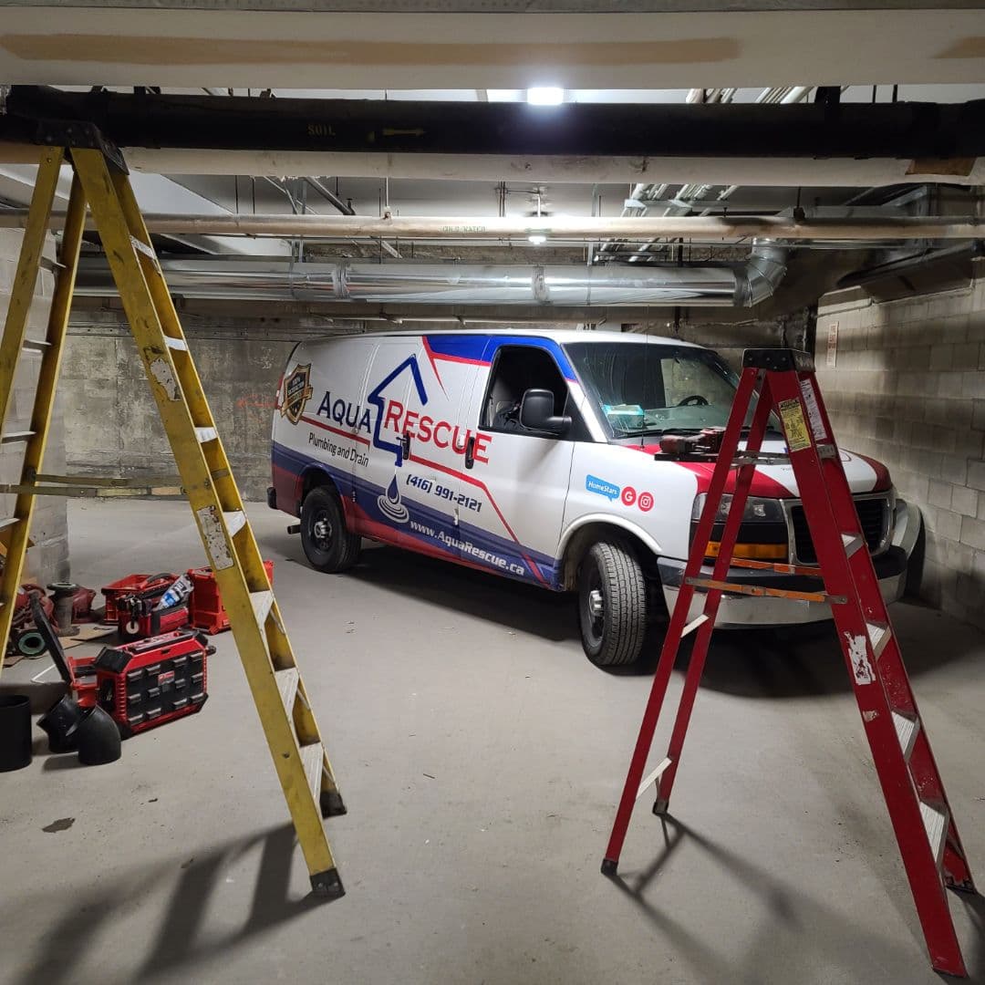 A white Aqua Rescue plumbing and drain service van is parked inside a concrete indoor garage. Two ladders, one yellow and one red, are positioned in the foreground on either side of the van. Various tools and toolboxes are scattered on the floor to the left of the vehicle. Overhead pipes and ductwork run along the ceiling. The scene is lit by ceiling lights.