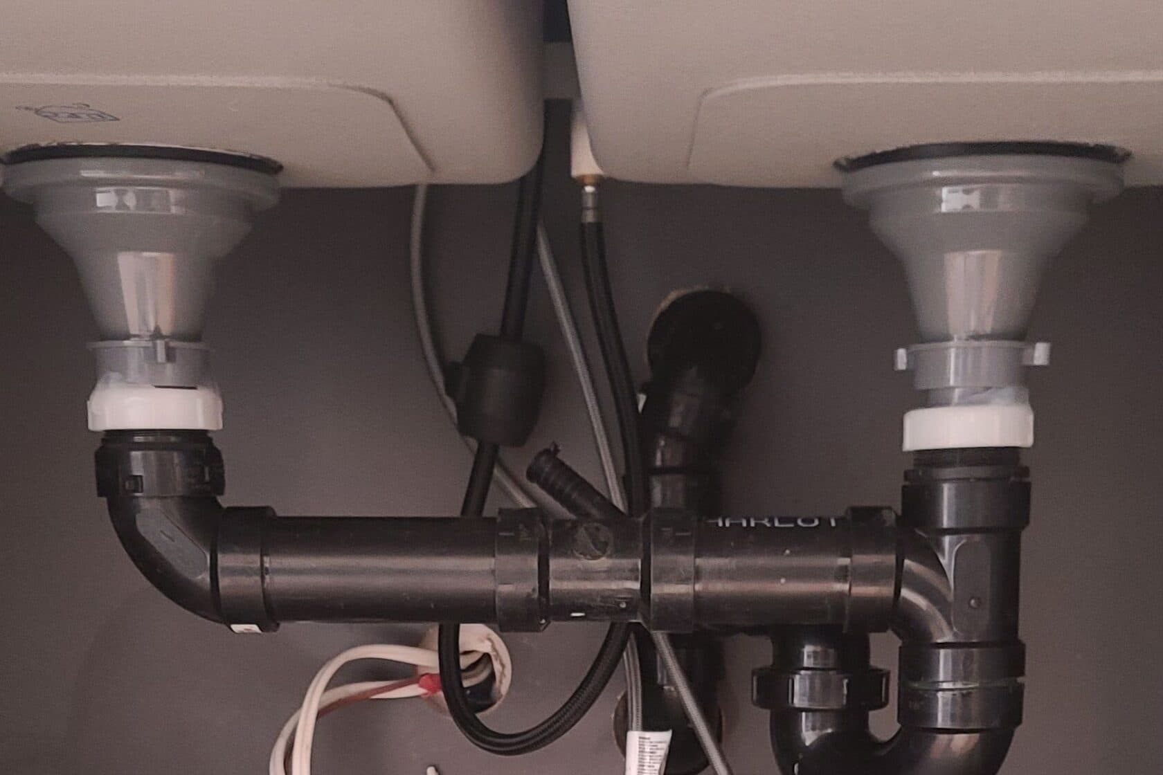 Under-sink view of a dual-basin kitchen sink plumbing setup with black PVC drain pipes and white compression fittings. The pipes connect both basins to a common trap and drain line, with electrical and water supply lines visible in the background.