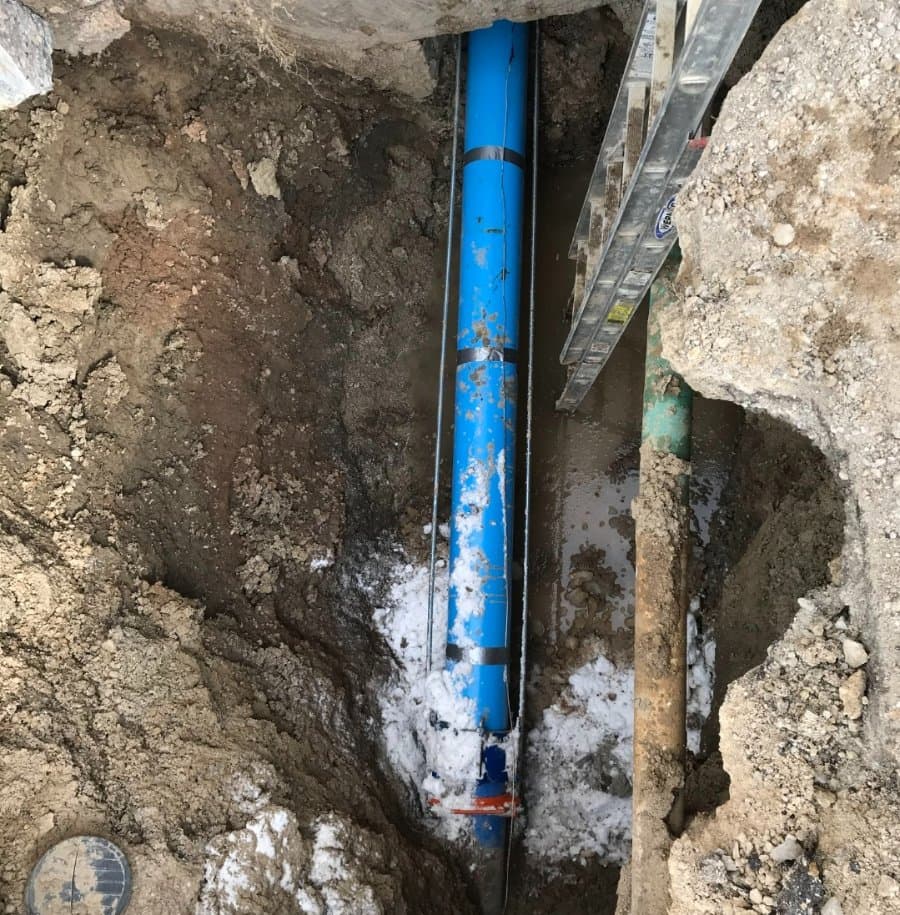 Underground trench showing new blue water main pipe installed beside an older green pipe, with ladder access and wet soil surrounding the excavation site.