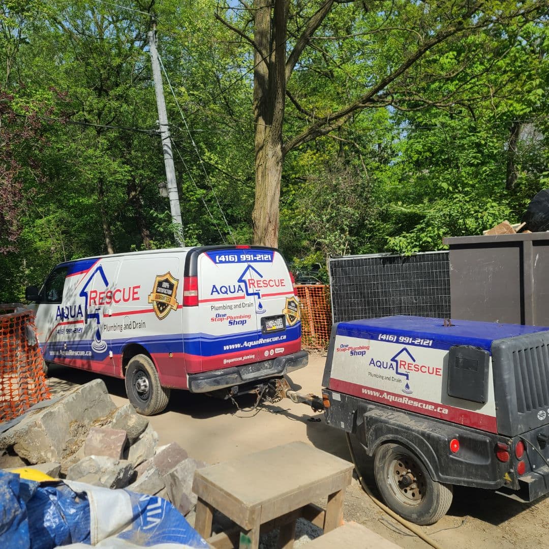 Aqua Rescue plumbing service van and matching utility trailer parked at a construction or repair site surrounded by trees and fencing. The van displays the company's branding, contact information, and tagline "Plumbing and Drain – One Stop Plumbing Shop." Construction materials, rocks, and equipment are visible around the site.