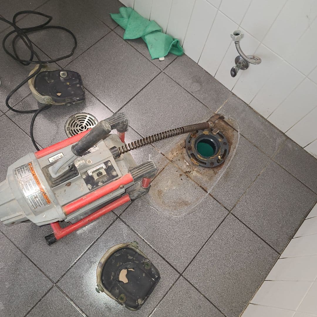 A drain snake machine is being used to unclog a toilet flange on a tiled bathroom floor. The toilet has been removed, exposing the drain opening, with the auger cable inserted for blockage removal. Nearby, toilet components and cleaning rags are visible, indicating active plumbing maintenance.