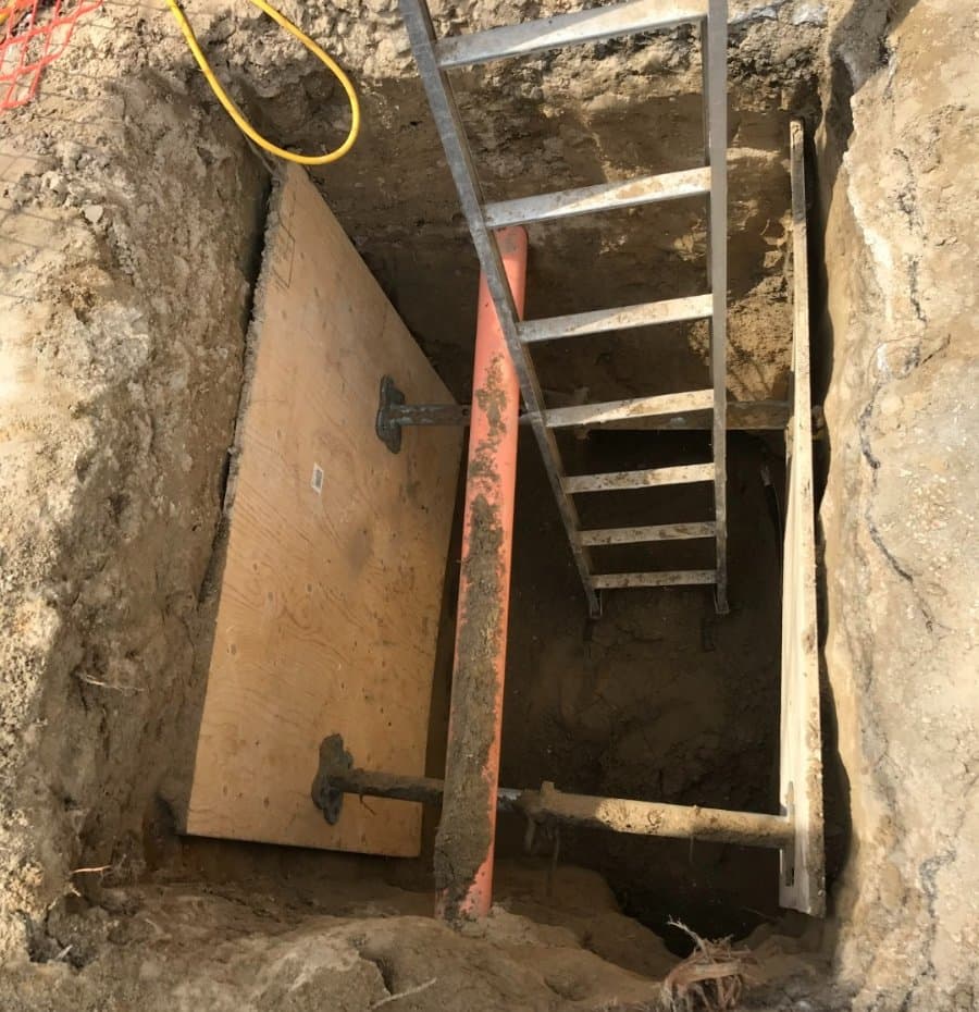 Excavated trench with trench box shoring system, metal ladder for access, and plywood panels for wall support, used for safe underground utility work.
