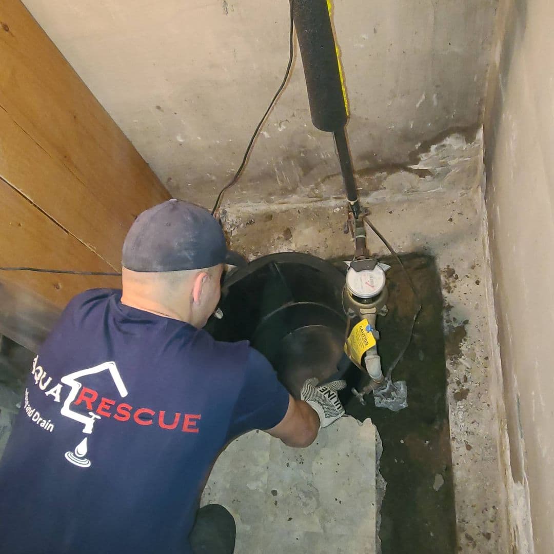 Technician from Aqua Rescue installing a sump pump basin in a basement corner with visible concrete floor and water meter setup nearby.
