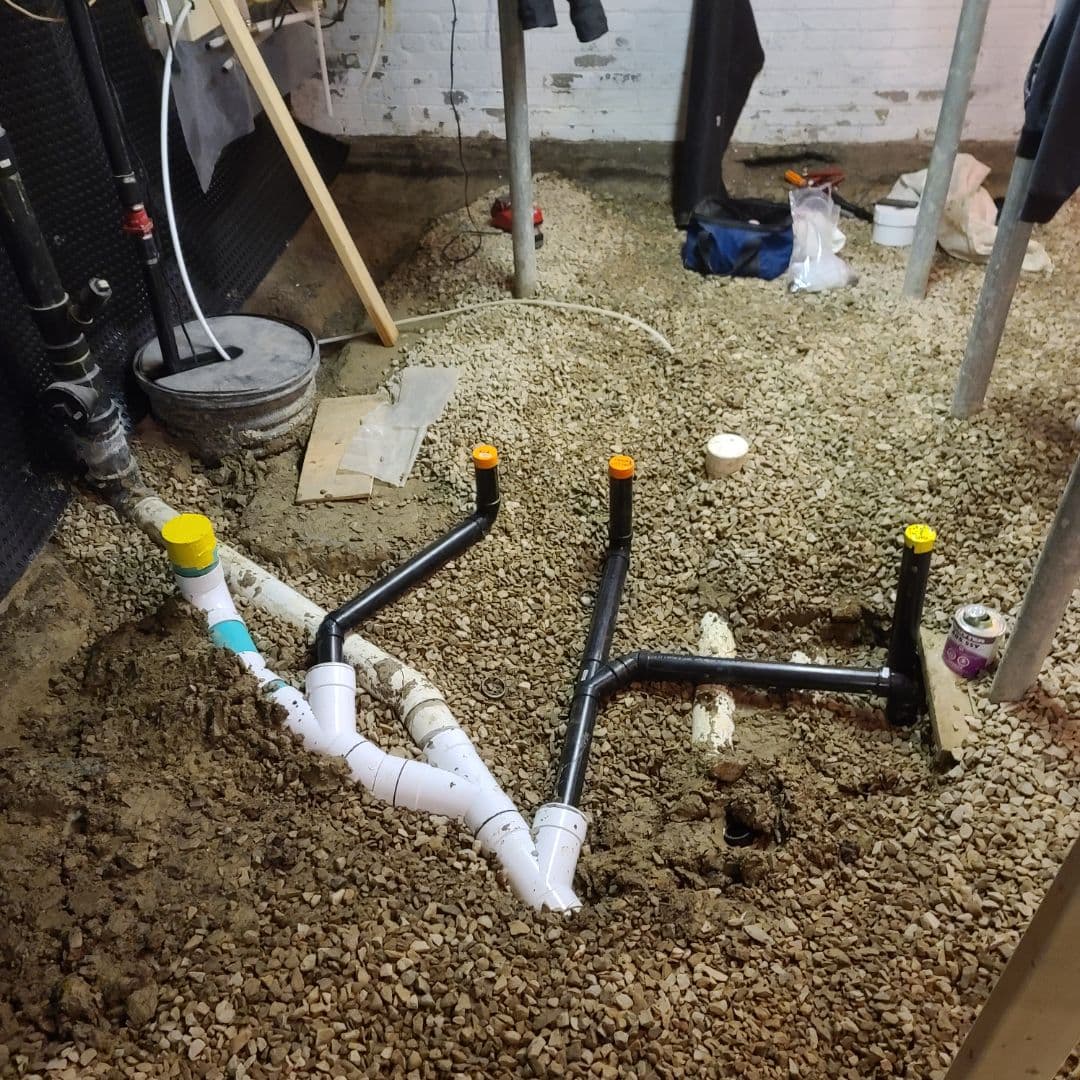 Basement plumbing installation in progress, showing newly laid black and white PVC pipes over a gravel bed. The pipes are capped and connected in preparation for drainage or sewage setup. Construction tools and materials are scattered around the unfinished basement area with concrete walls and support columns.