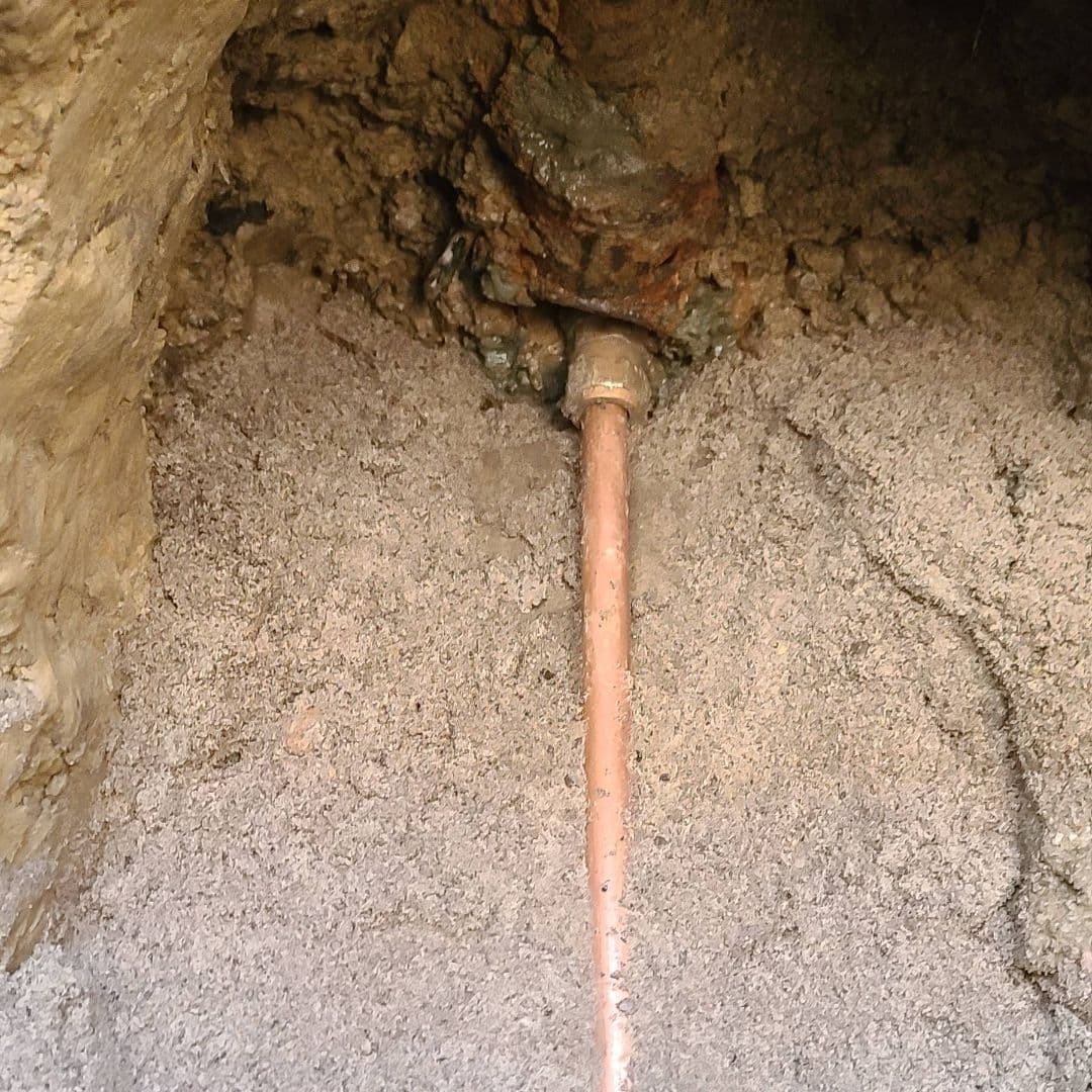 A close-up view of an exposed copper water pipe emerging from a dirt wall, connected to an older, corroded underground fitting. The surrounding area appears to be part of an excavation site, revealing the connection for potential repair or replacement.