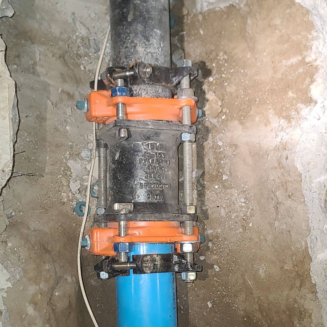 A close-up image of a pipe repair using a mechanical coupling connecting a black metal pipe to a blue plastic pipe. The joint is reinforced with orange and silver metal clamps, surrounded by a concrete surface.