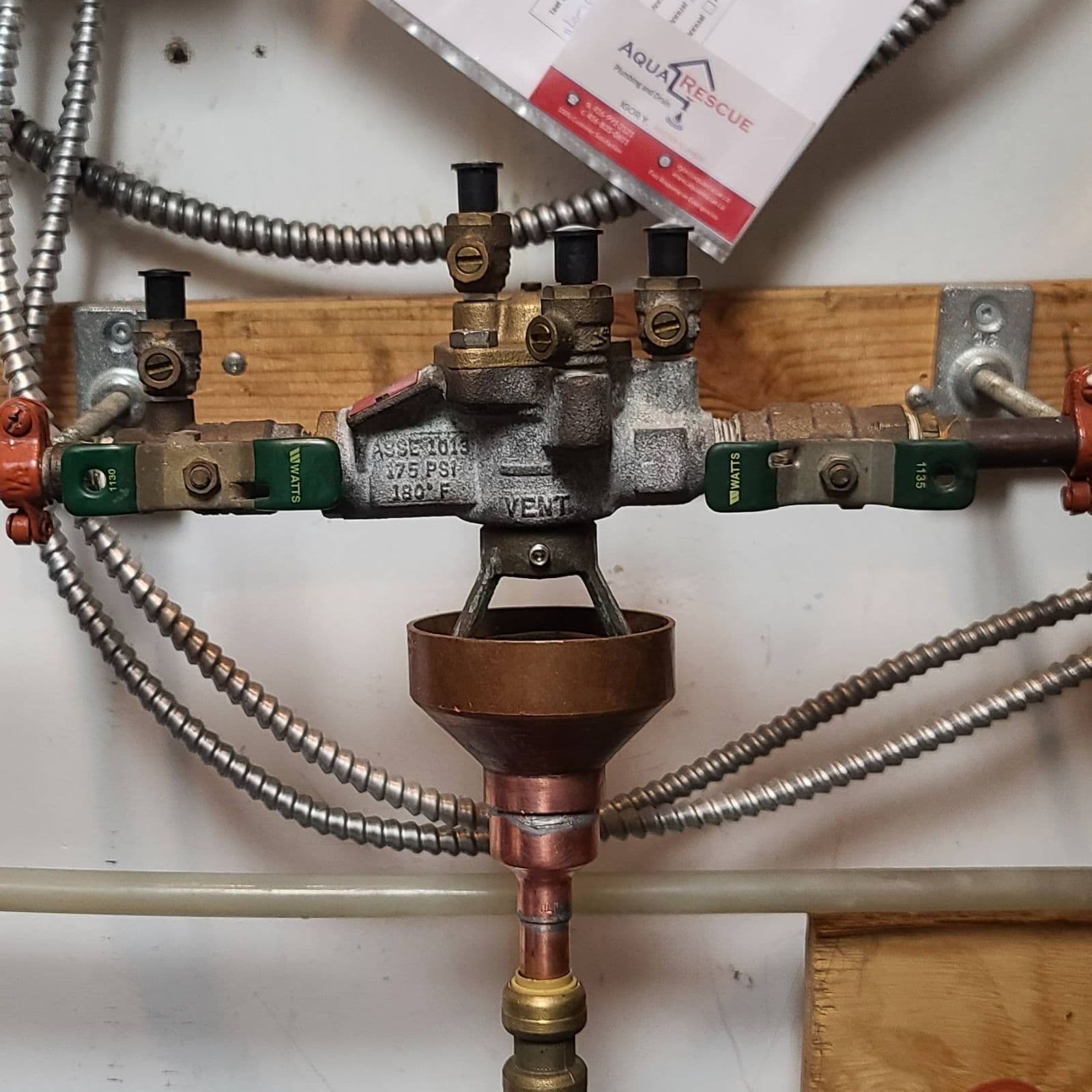 Close-up of a backflow preventer assembly installed on a wall-mounted plumbing system, featuring multiple brass valves with green handles, black test ports, and a central vent. A copper funnel-shaped drain is attached below the device, with flexible metal conduit and water lines surrounding the setup. An Aqua Rescue service sheet is partially visible at the top of the image.
