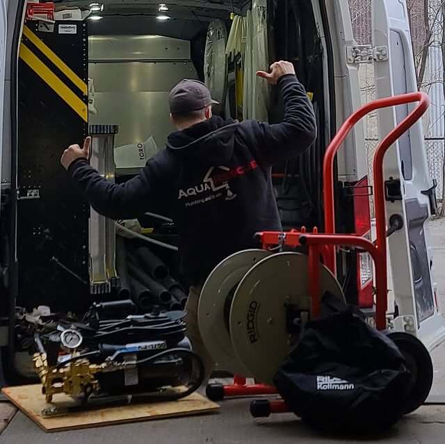 A worker wearing a black Aqua Rescue hoodie stands at the back of a service van, surrounded by plumbing equipment including a hose reel, pipes, and a pump, preparing for a plumbing job.