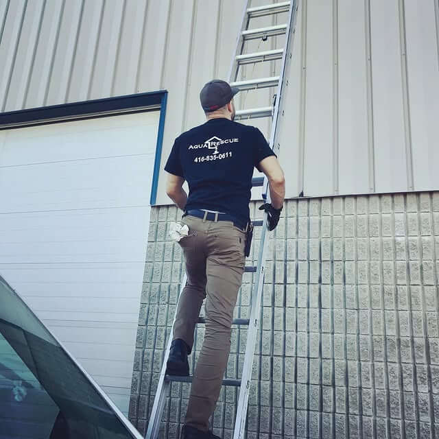 Person wearing an Aqua Rescue t-shirt and cap climbing a tall aluminum ladder leaning against the side of a light-colored industrial building with a roll-up garage door. Part of a car is visible in the foreground.