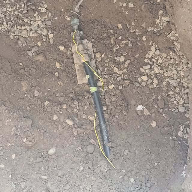 A section of underground piping partially exposed in a dirt trench, with small gravel scattered around. The pipe is wrapped in black protective tape and has a yellow grounding wire attached, secured with tape.