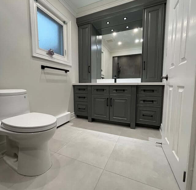 A modern bathroom with a white toilet on the left and a gray vanity with multiple drawers and cabinet doors on the right. Above the vanity is a large mirror with built-in lights, and the floor is covered with large light gray tiles. A frosted window is on the wall above a black towel bar. The overall color scheme is neutral with white walls and gray accents.