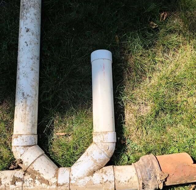 A section of plumbing pipes laid on grass. The setup includes white PVC pipes and an older, discolored clay or terracotta pipe. The pipes form a T-junction, with the PVC pipe extending upward from the joint. Dirt is visible on parts of the pipes.