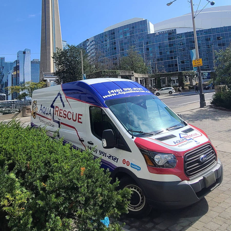 Aqua Rescue Plumbing and Drain service van parked on a city sidewalk in front of the CN Tower and the Rogers Centre in downtown Toronto. The van is white with red and blue branding, including contact information and website on the side and windshield. Surrounding the van are bushes, a city street, and modern high-rise buildings.