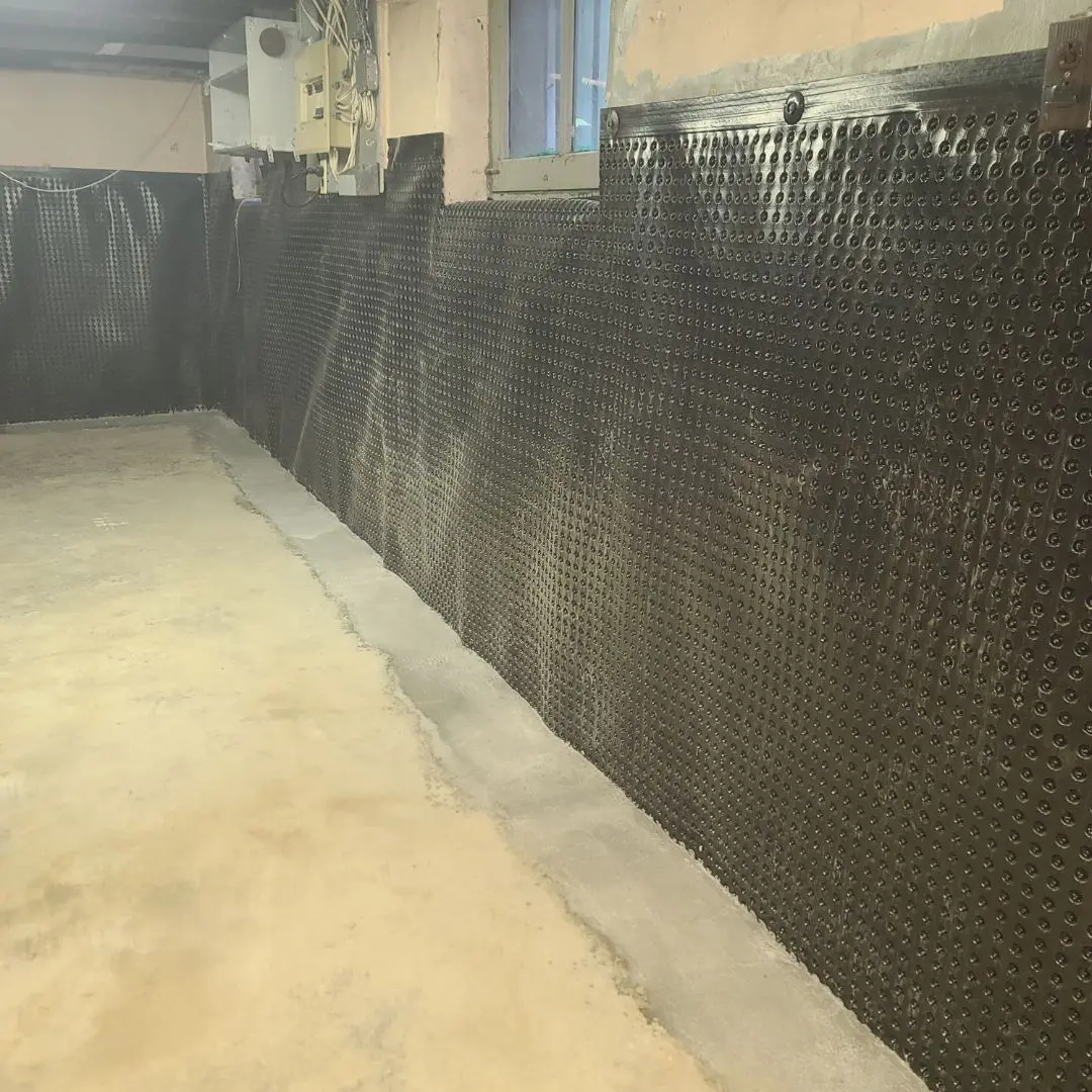 Interior basement wall lined with dimpled waterproof membrane and a fresh layer of concrete along the base for foundation drainage and water intrusion prevention.