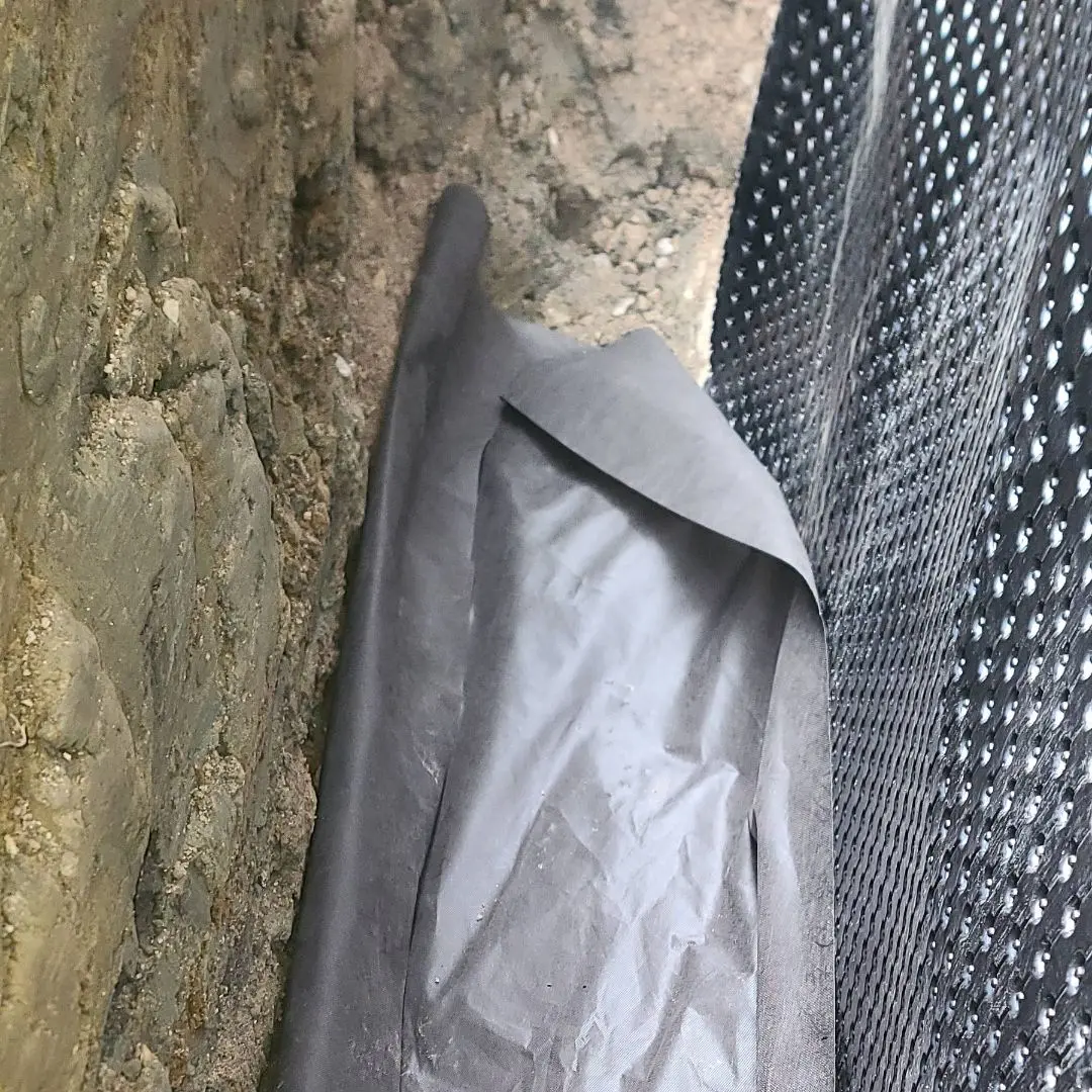 Close-up of a foundation wall with waterproofing membrane and geotextile fabric lining a trench for exterior drainage installation.