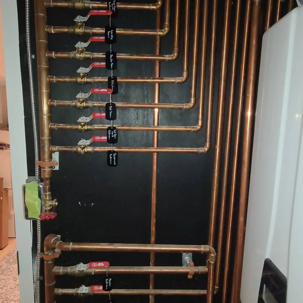 A neatly organized plumbing manifold with multiple copper pipes mounted vertically on a black wall. Each pipe has a red-handled shut-off valve labeled with a small black tag, indicating areas like “Laundry,” “Bathroom,” and “Kitchen.” Two horizontal copper pipes run across the bottom, also equipped with red-handled valves and labeled tags. The installation appears clean and precise, with smooth solder joints and consistent spacing.
