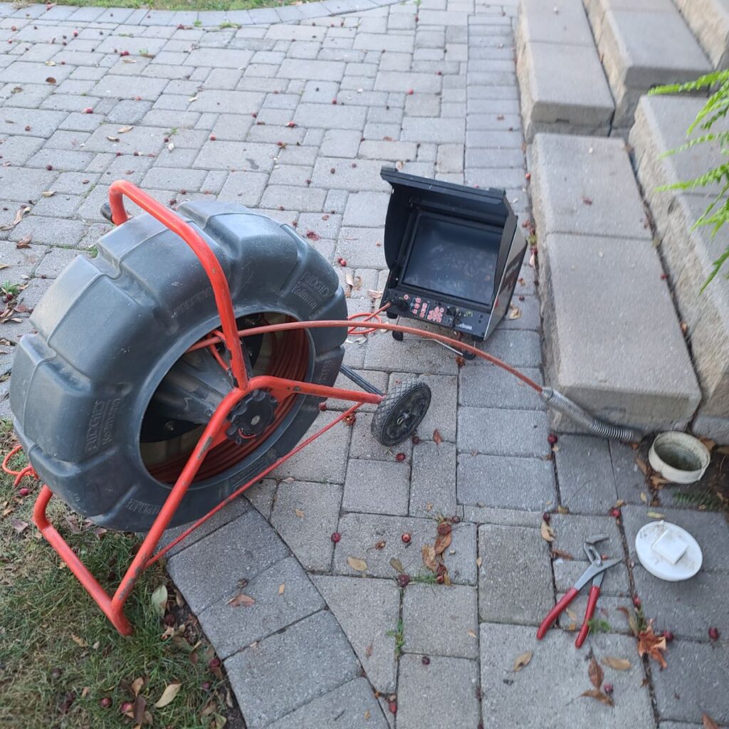 A drain inspection camera setup on a paved walkway next to stone steps. The device has a large black cable reel with a red frame, a flexible camera cable inserted into a white floor drain, and a small monitor resting on the ground. A pair of pliers lies nearby, and a few leaves and berries are scattered on the pavers.