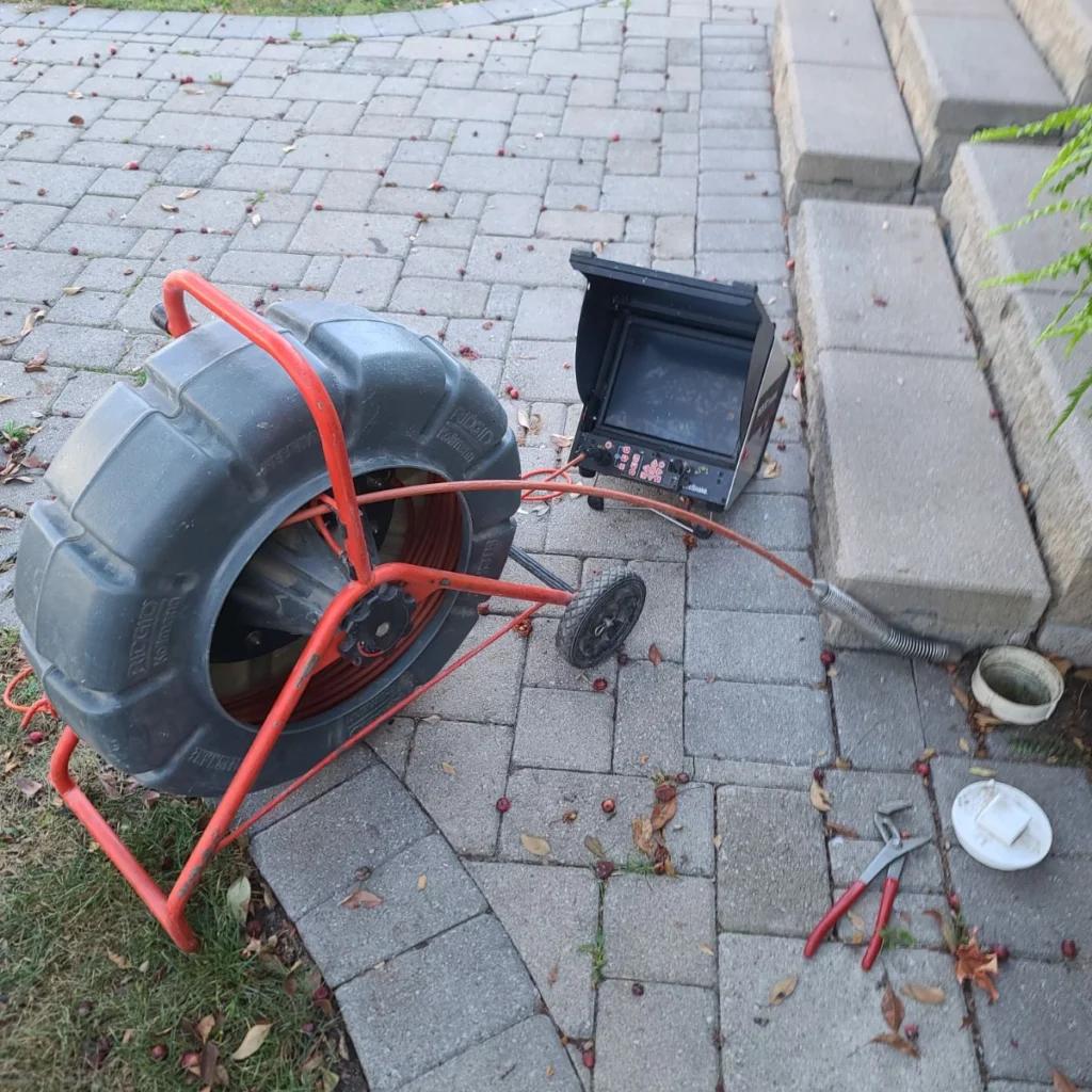 A drain inspection camera setup on a paved walkway next to stone steps. The device has a large black cable reel with a red frame, a flexible camera cable inserted into a white floor drain, and a small monitor resting on the ground. A pair of pliers lies nearby, and a few leaves and berries are scattered on the pavers.