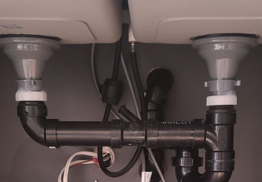 Under-sink view of a dual-basin kitchen sink plumbing setup with black PVC drain pipes and white compression fittings. The pipes connect both basins to a common trap and drain line, with electrical and water supply lines visible in the background.
