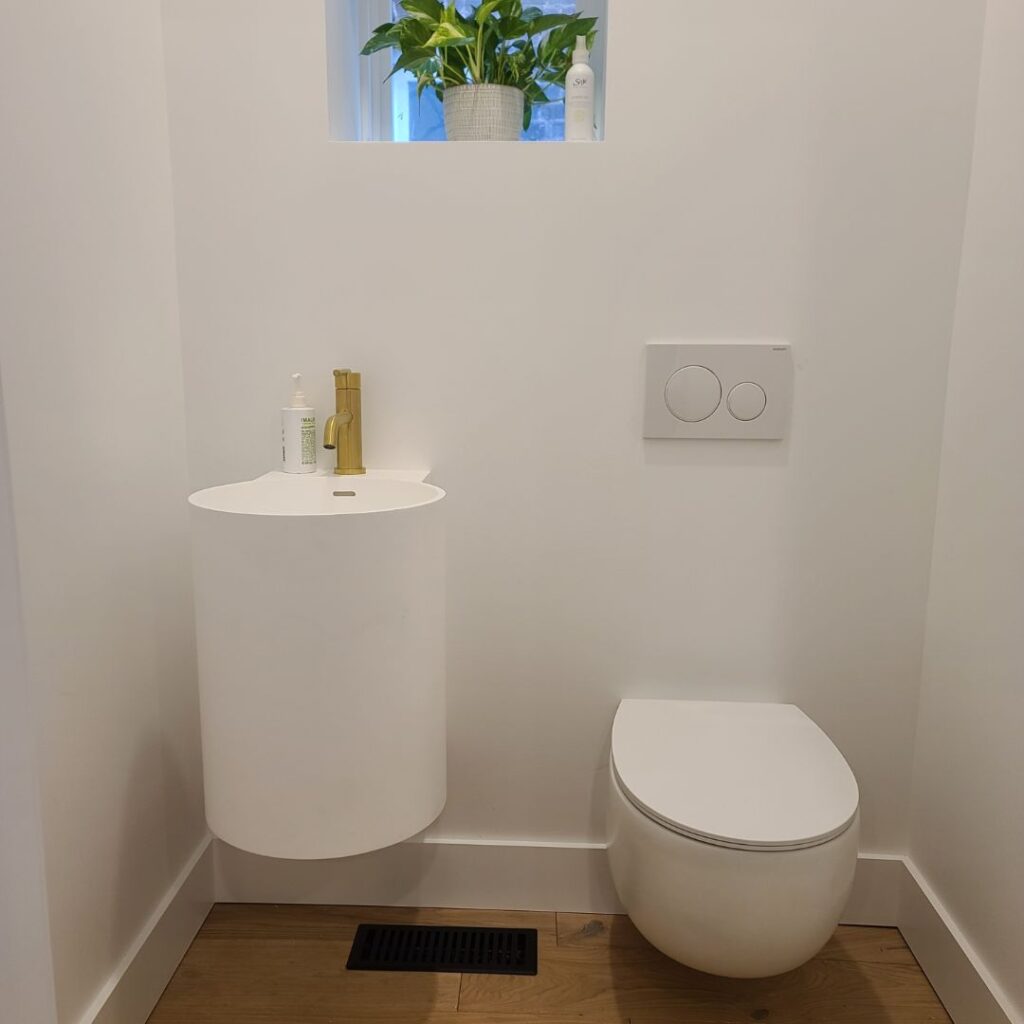 Minimalist modern bathroom with a wall-mounted white toilet and a compact white cylindrical sink featuring a brass faucet. A small window above the sink holds a potted plant and toiletry items, adding a touch of greenery to the clean, white space. The floor is finished with light wood and a black floor vent is visible beneath the sink.