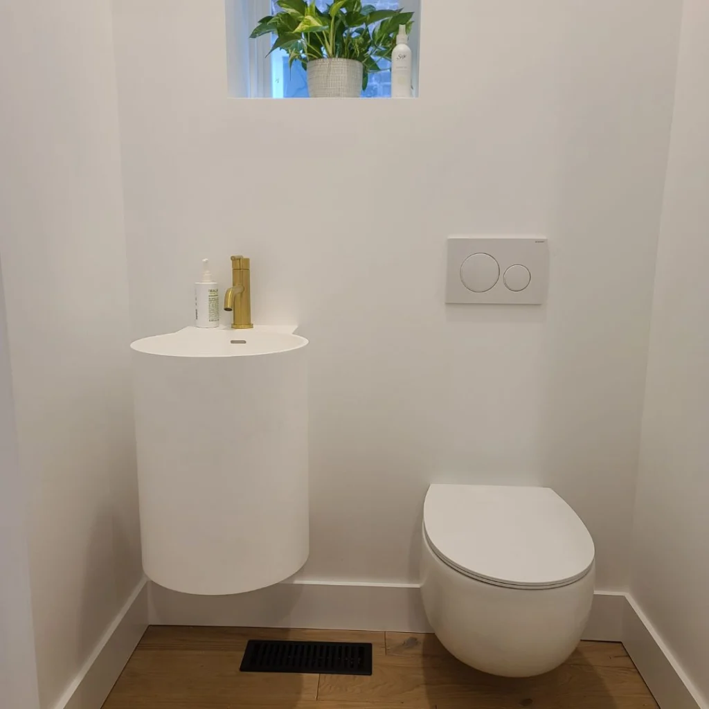 Minimalist modern bathroom with a wall-mounted white toilet and a compact white cylindrical sink featuring a brass faucet. A small window above the sink holds a potted plant and toiletry items, adding a touch of greenery to the clean, white space. The floor is finished with light wood and a black floor vent is visible beneath the sink.