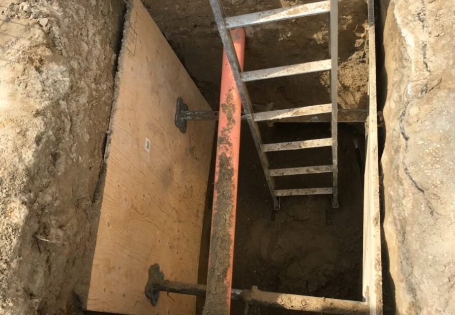 Excavated trench with trench box shoring system, metal ladder for access, and plywood panels for wall support, used for safe underground utility work.