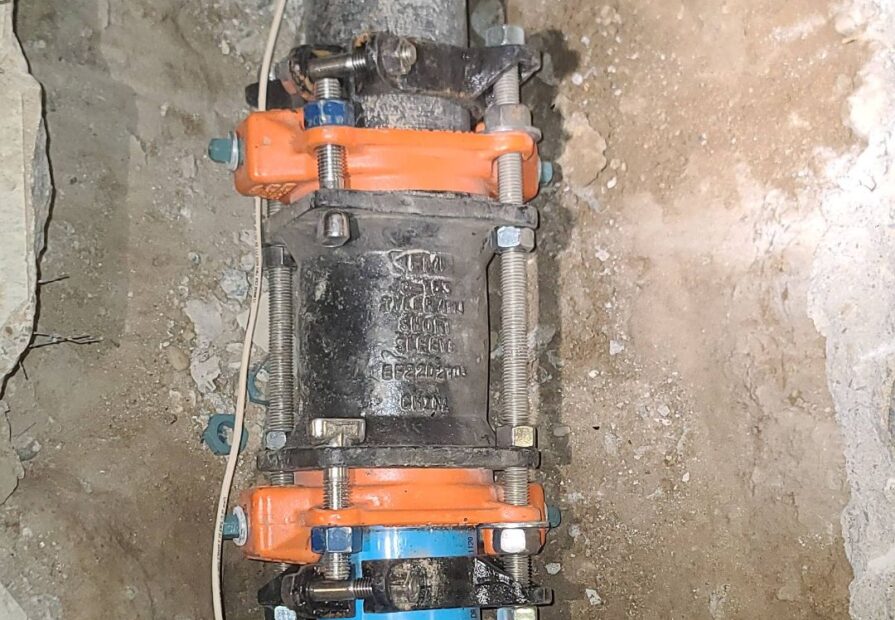 A close-up image of a pipe repair using a mechanical coupling connecting a black metal pipe to a blue plastic pipe. The joint is reinforced with orange and silver metal clamps, surrounded by a concrete surface.