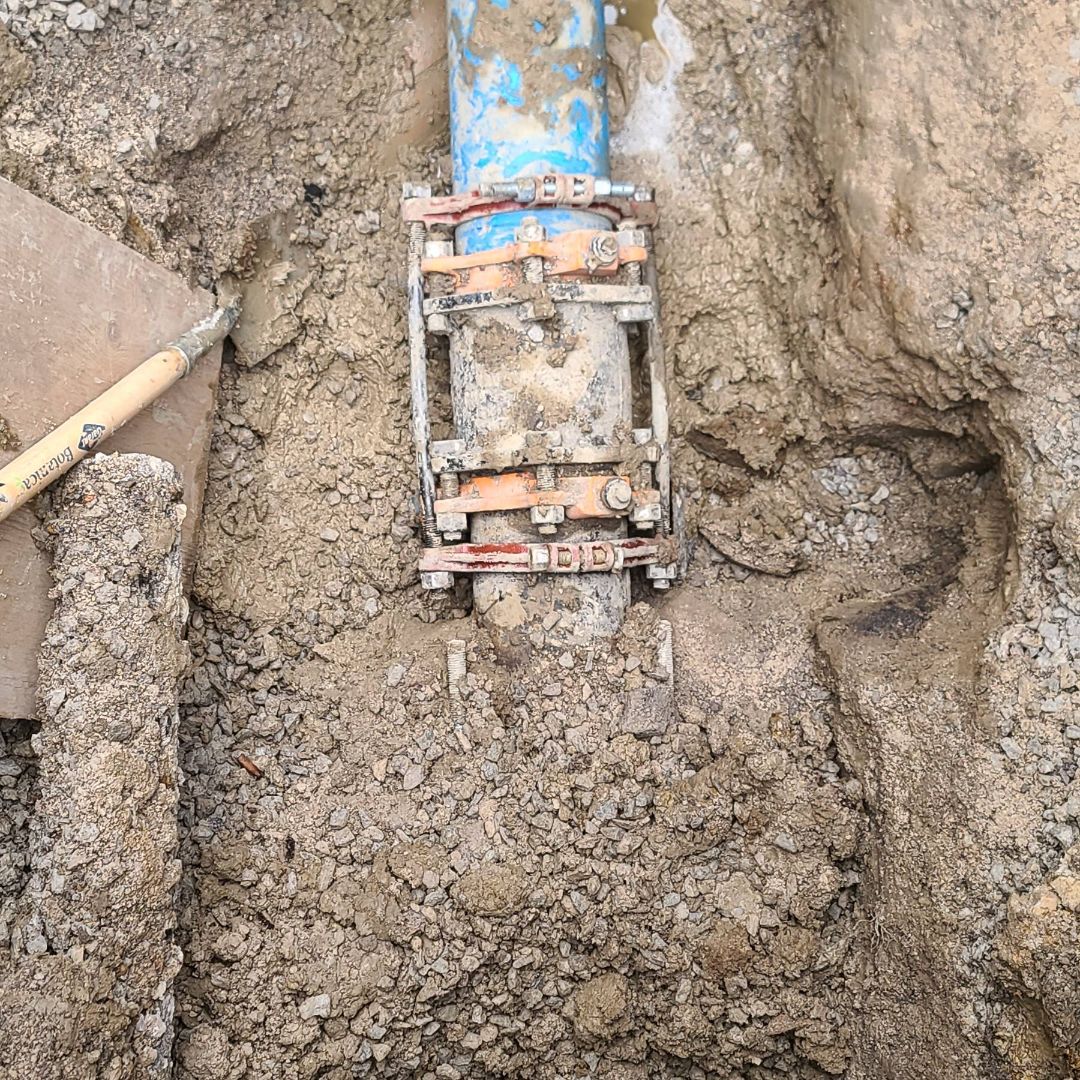 Close-up of an underground water pipe with a metal repair clamp, partially buried in soil, next to a shovel and digging tools.