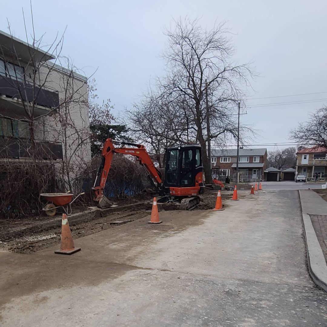 Small excavator surrounded by traffic cones working on a residential street construction site near apartment buildings and houses.